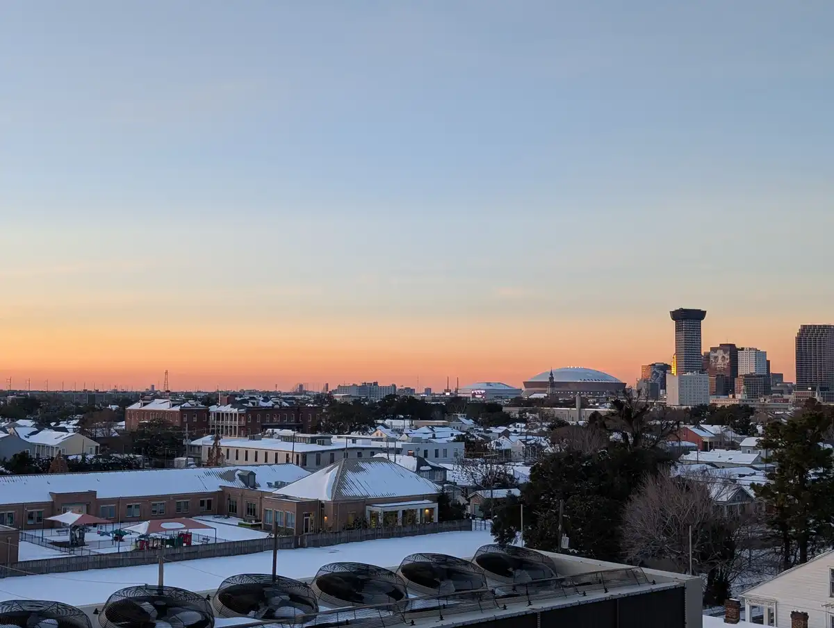 A scenic view of a snow-covered New Orleans cityscape at sunset, featuring rooftops blanketed in white, the Superdome, and…