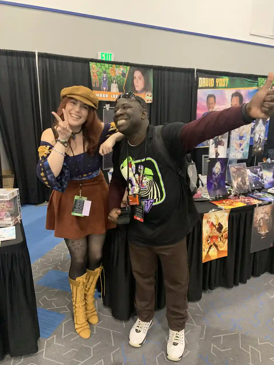 Two people are posing for a photo at a convention booth. The woman on the left, wearing a brown beret, a star-patterned top…