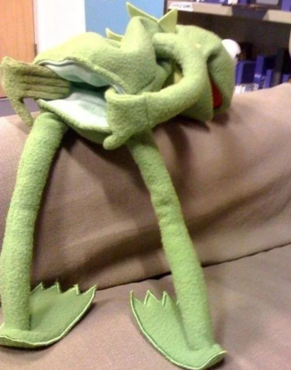 A green felt Kermit the Frog plush toy is sitting on a brown couch, hugging itself tightly with both arms wrapped around its…
