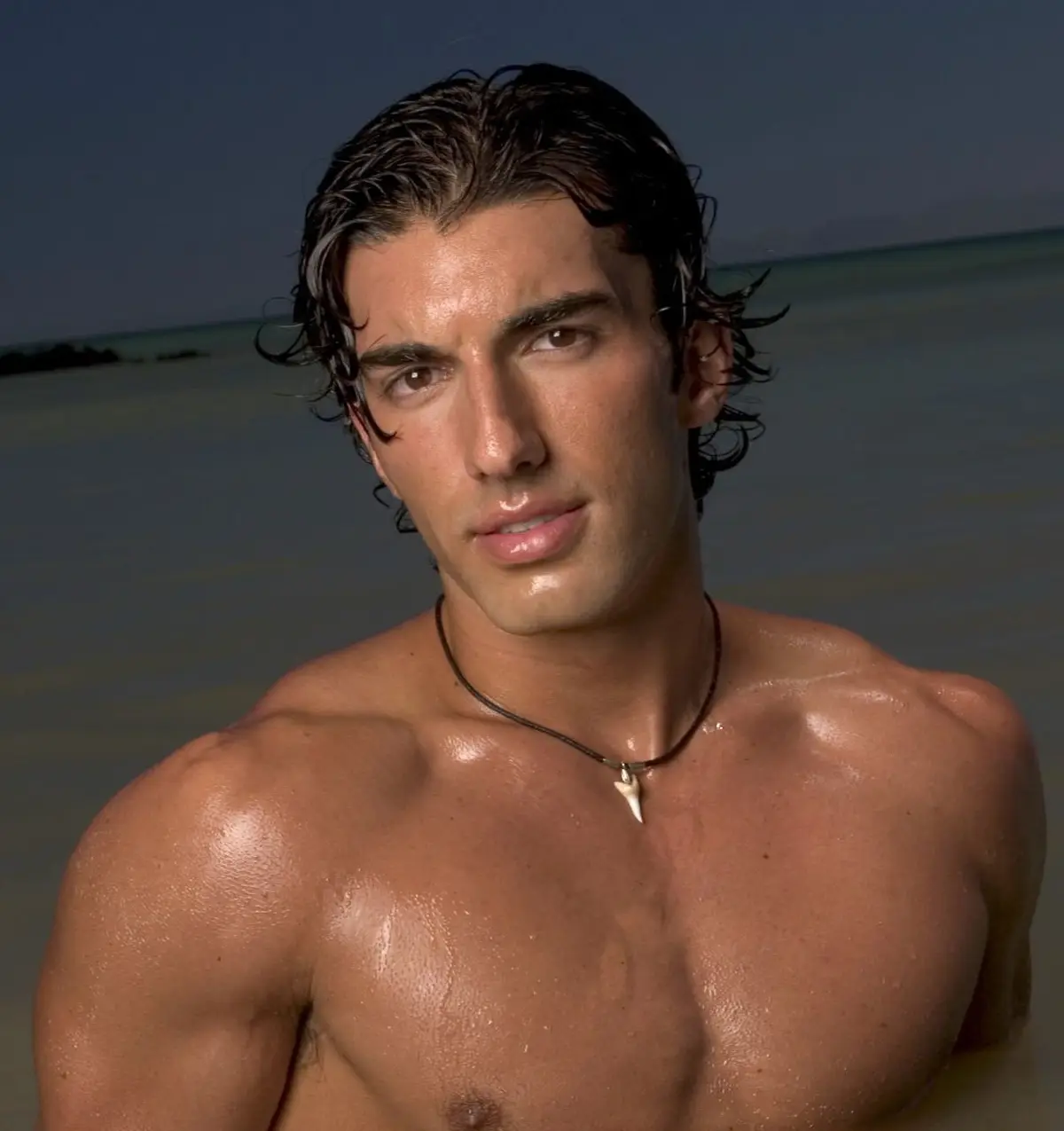A shirtless, muscular man with wet, dark hair stands in shallow ocean water, looking directly at the camera with a serious…