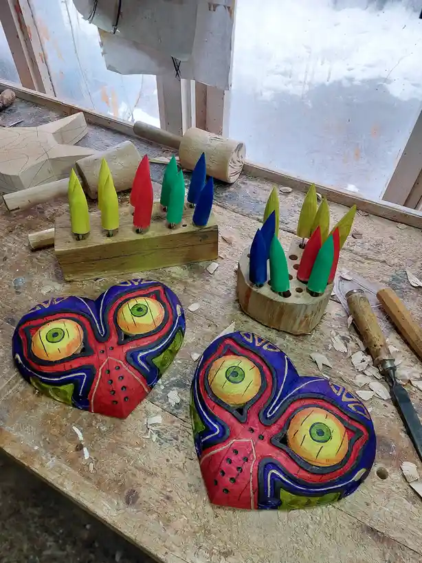 Two hand-carved wooden masks with large, colorful eyes and intricate patterns sit on a workshop table surrounded by wood…