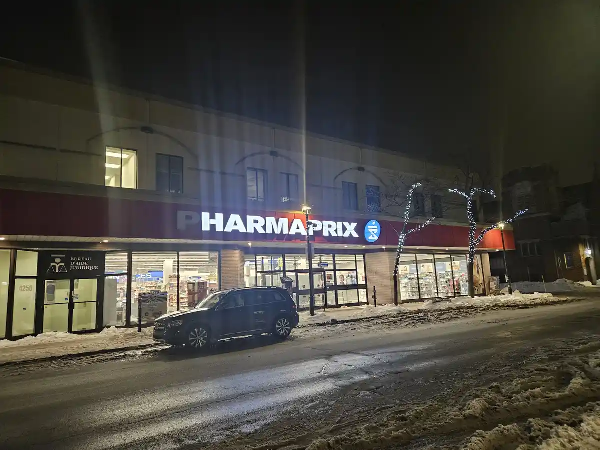 A nighttime photo of a Pharmaprix pharmacy storefront with snow on the ground and a dark SUV parked in front. The building…