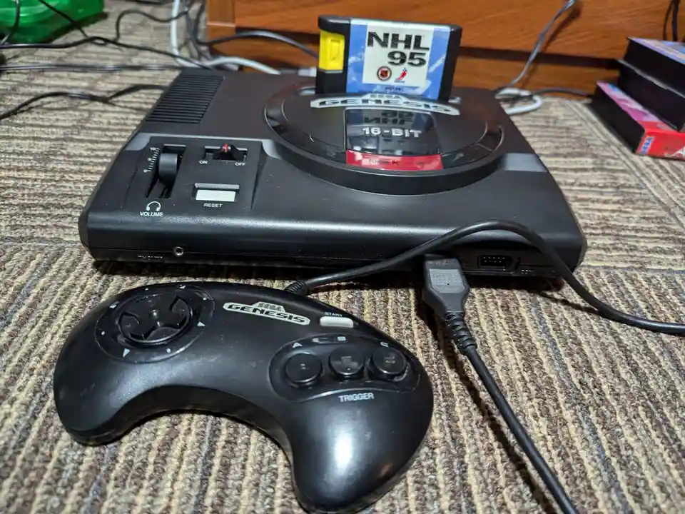 A black Sega Genesis console sits on a striped carpet with a 'NHL 95' game cartridge inserted into its slot. In front of it…
