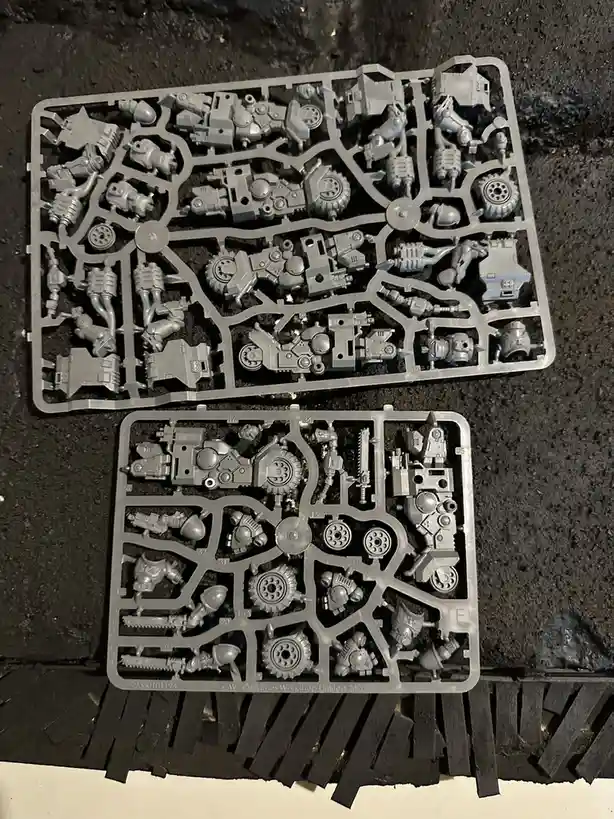 Two gray plastic sprues filled with unpainted miniature parts for a tabletop wargame lie on a dark, textured surface. The…