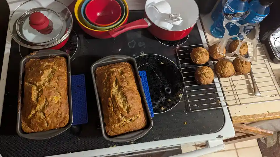 An overhead photo shows a cluttered stovetop with two loaves of banana bread in metal pans, a cooling rack with muffins, and…