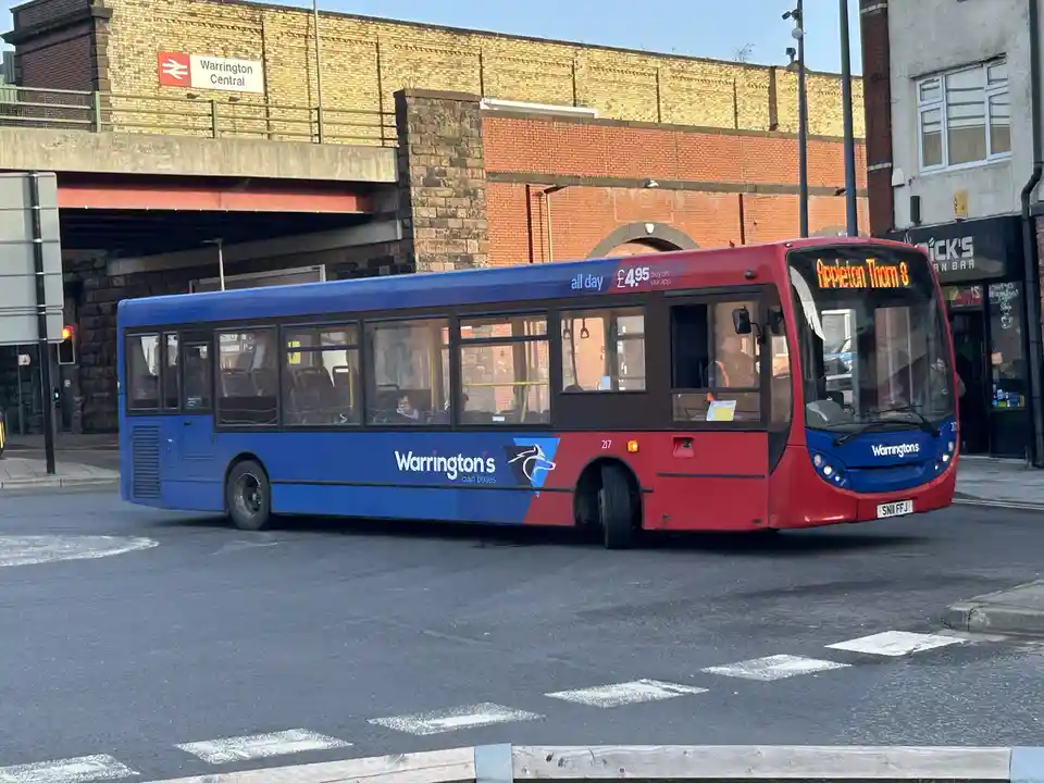 A blue and red Warrington's Own Buses vehicle, number 217, is shown on a street near Warrington Central railway station. The…