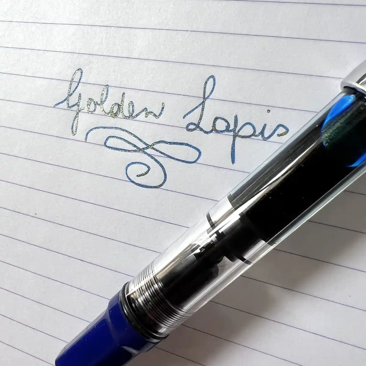 A close-up photo shows a blue fountain pen resting on lined notebook paper, with the words 'golden lapis' written in cursive…
