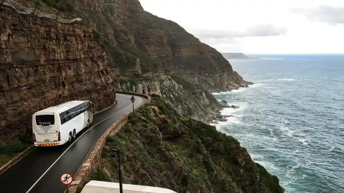 Winding road hugs the rugged coastline, where turquoise waves crash against ancient cliffs, their rust-red hues glowing in…