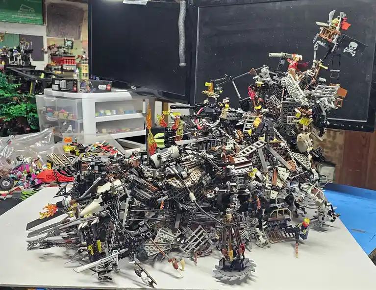 A massive, chaotic pile of LEGO pieces and minifigures, resembling a wrecked pirate ship or war machine, sits on a white…
