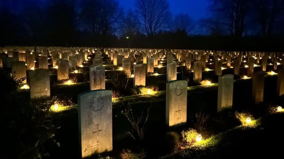 A nighttime photograph of a vast cemetery with rows of uniform headstones, each illuminated by a small candle at its base…