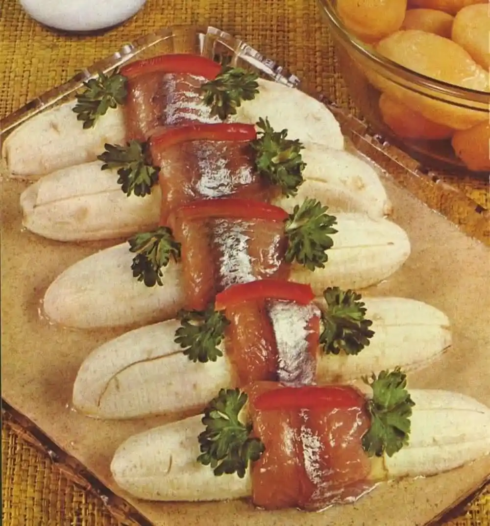 A vintage food photograph shows several peeled bananas arranged on a glass plate, each topped with a small, whole sardine…