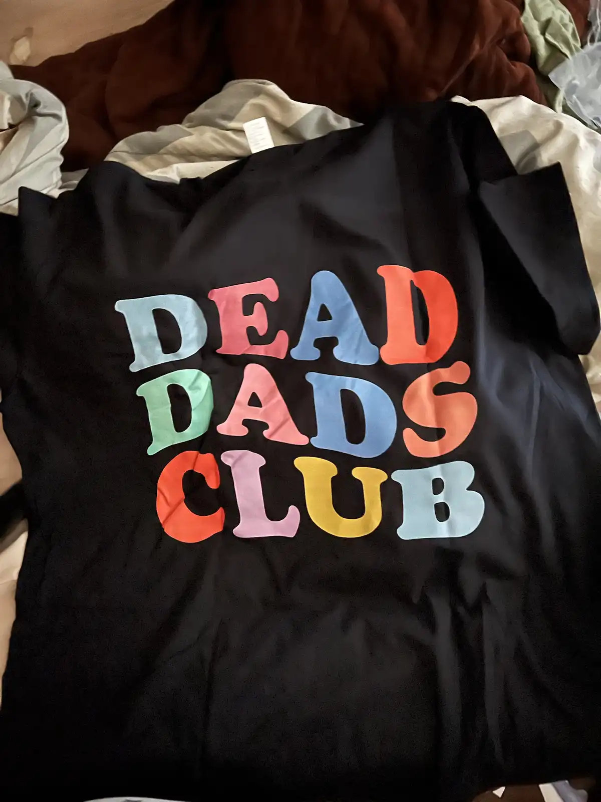 A black t-shirt laid on rumpled bedding displays the phrase 'DEAD DADS CLUB' in large, colorful, blocky letters with each…
