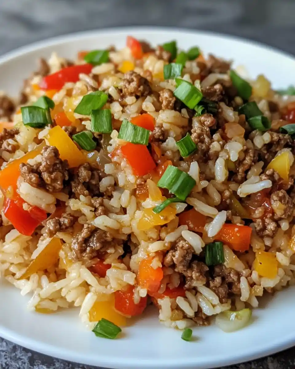 a close-up photo of a white plate filled with a colorful rice dish containing brown rice, ground meat, diced red and yellow…