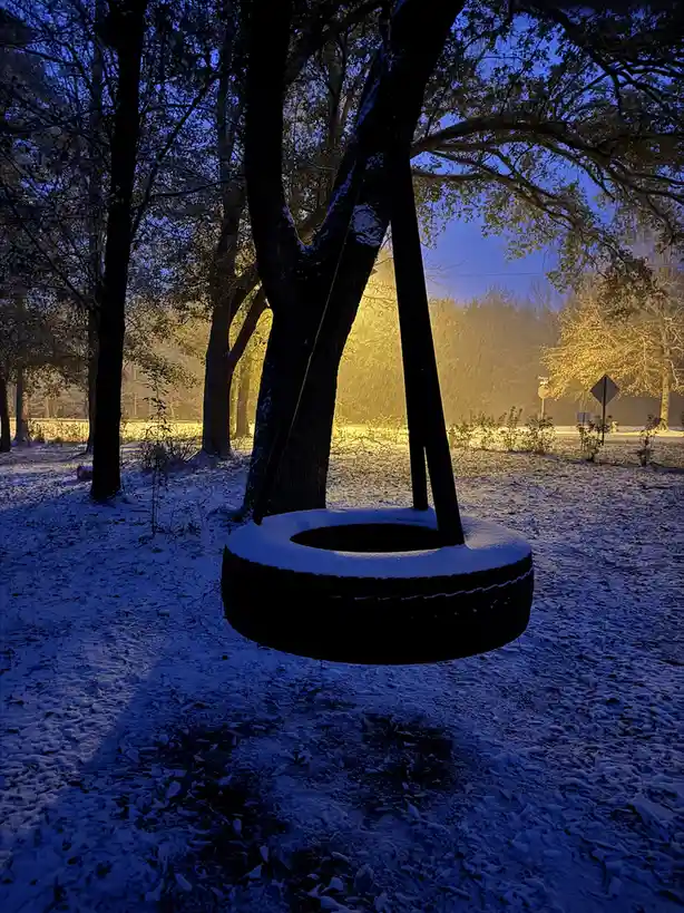 A snow-covered tire swing hangs from a tree in a snowy park at dusk, with a warm, golden light illuminating the background…