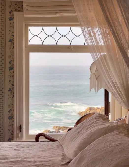A serene bedroom view from a bed with white linens, looking out a window with decorative glass panes at a rocky coastline…