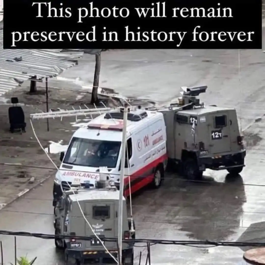 An aerial view shows a white ambulance with red stripes and Arabic/English text parked next to two armored military vehicles…