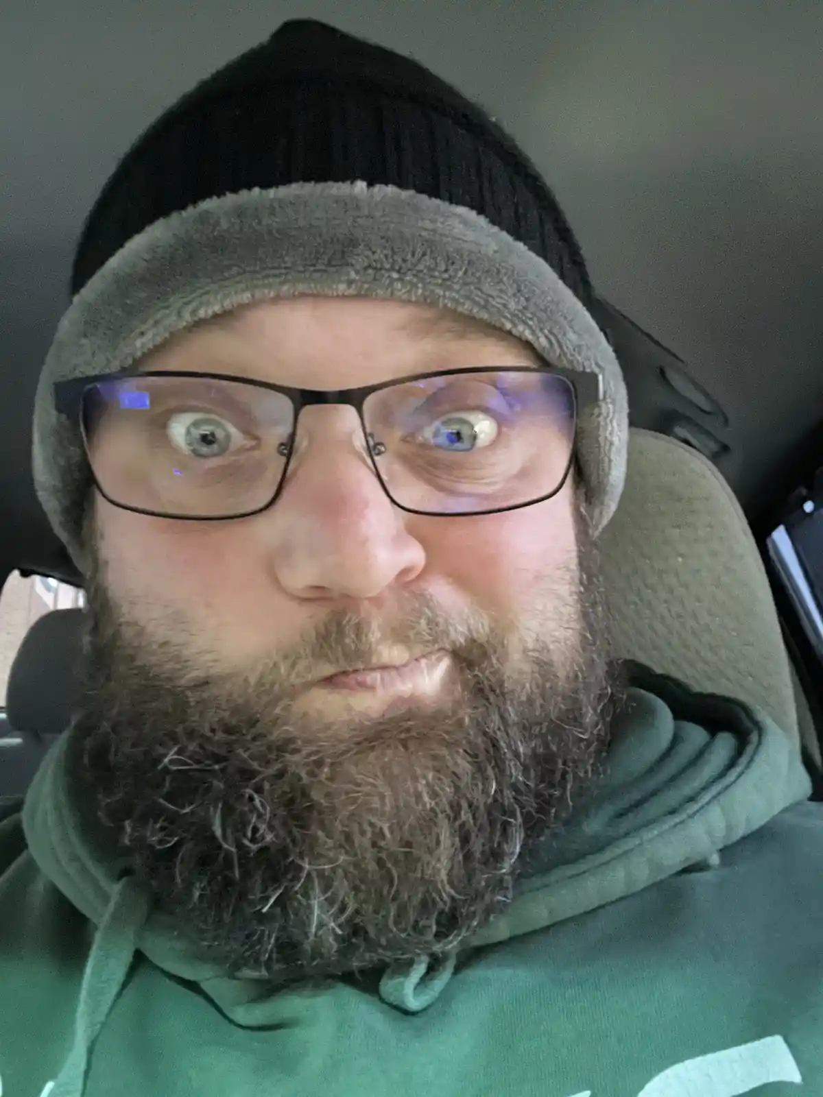 A man with a beard, glasses, and a black beanie with a grey fleece trim sits in a car seat, staring directly at the camera…