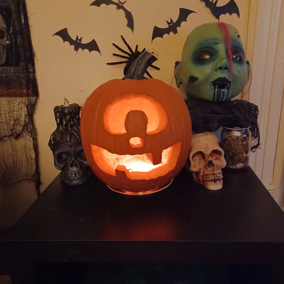 A carved, glowing jack-o'-lantern with a face shaped like a question mark sits on a black table surrounded by Halloween…