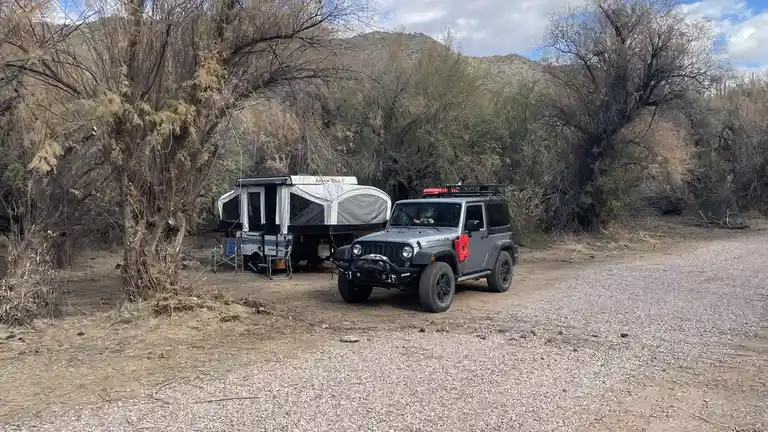 A silver Jeep Wrangler with a roof rack and red fuel canister is parked on a gravel road next to a white pop-up camper…