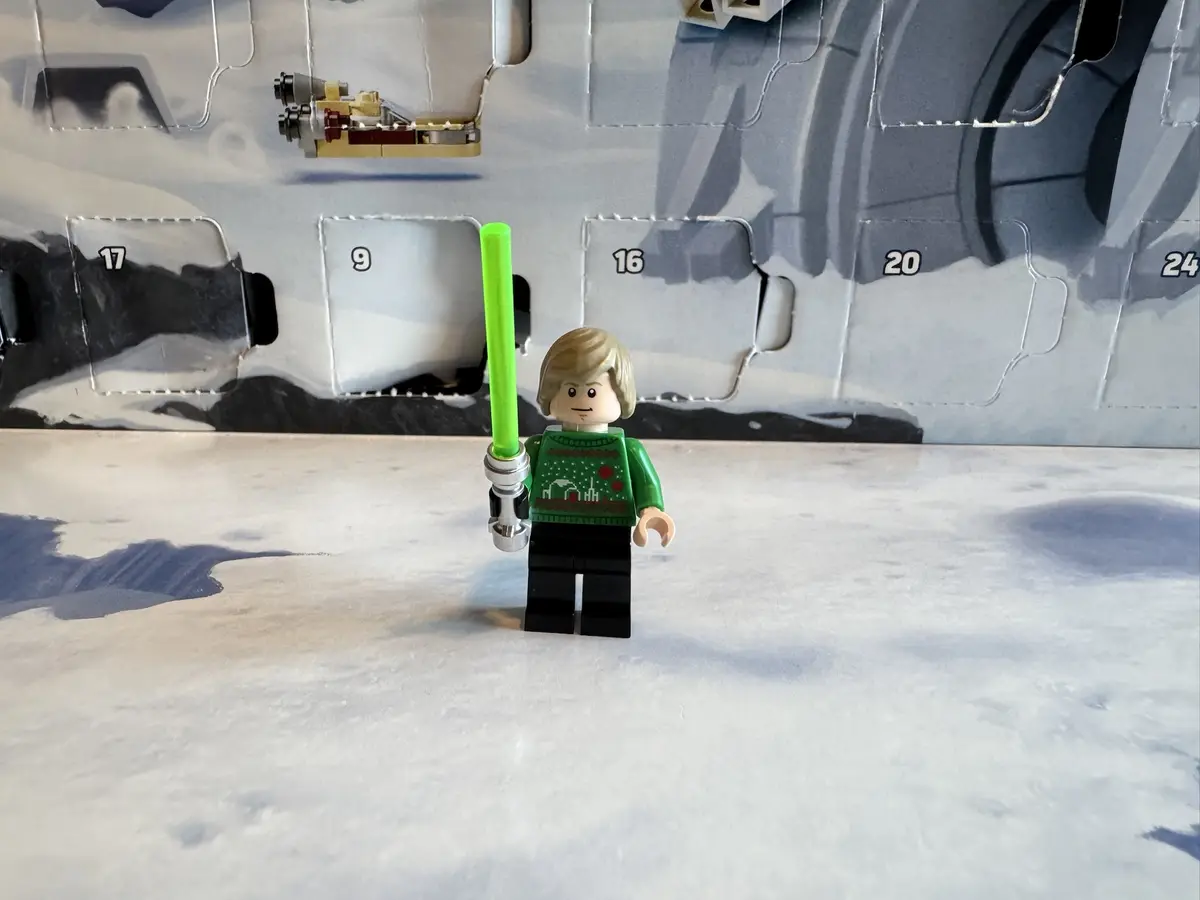A Lego minifigure with blonde hair, wearing a green Christmas sweater with red and white patterns, stands in front of a…