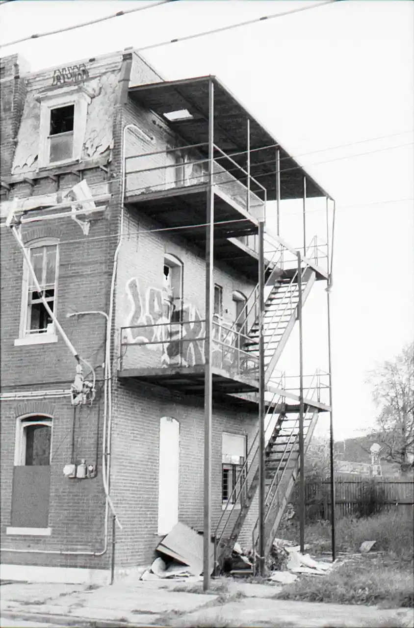 A black-and-white photo of a dilapidated brick building with a fire escape, broken windows, graffiti, and debris. The…