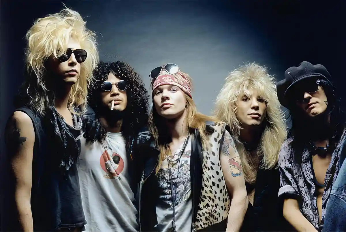 A promotional photo of the rock band Guns N' Roses, featuring five members with long hair, sunglasses, and tattoos, posing…