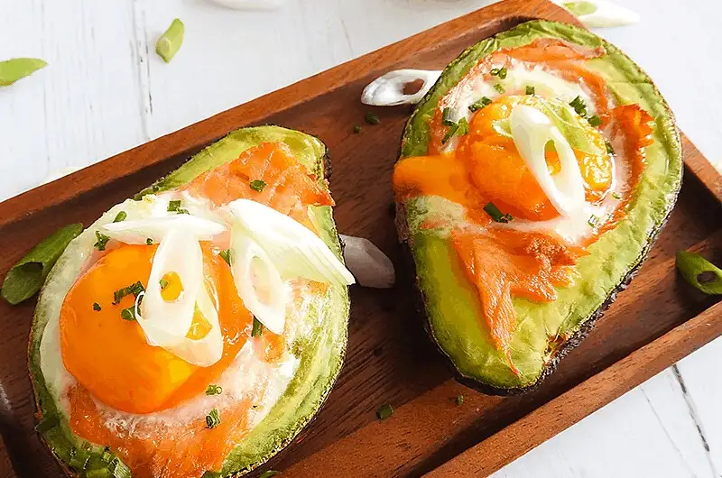 Two avocado halves on a wooden board, each filled with a fried egg, slices of salmon, and garnished with chopped chives and…