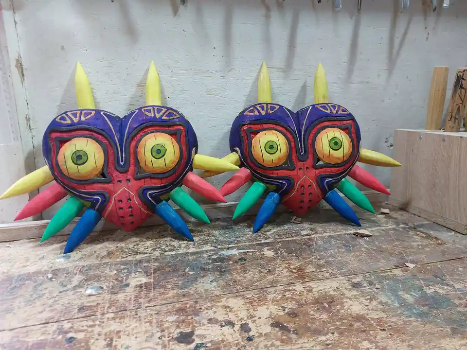 Two hand-carved wooden masks resembling Majora's Mask from The Legend of Zelda, with colorful spikes and large eyes, sit…