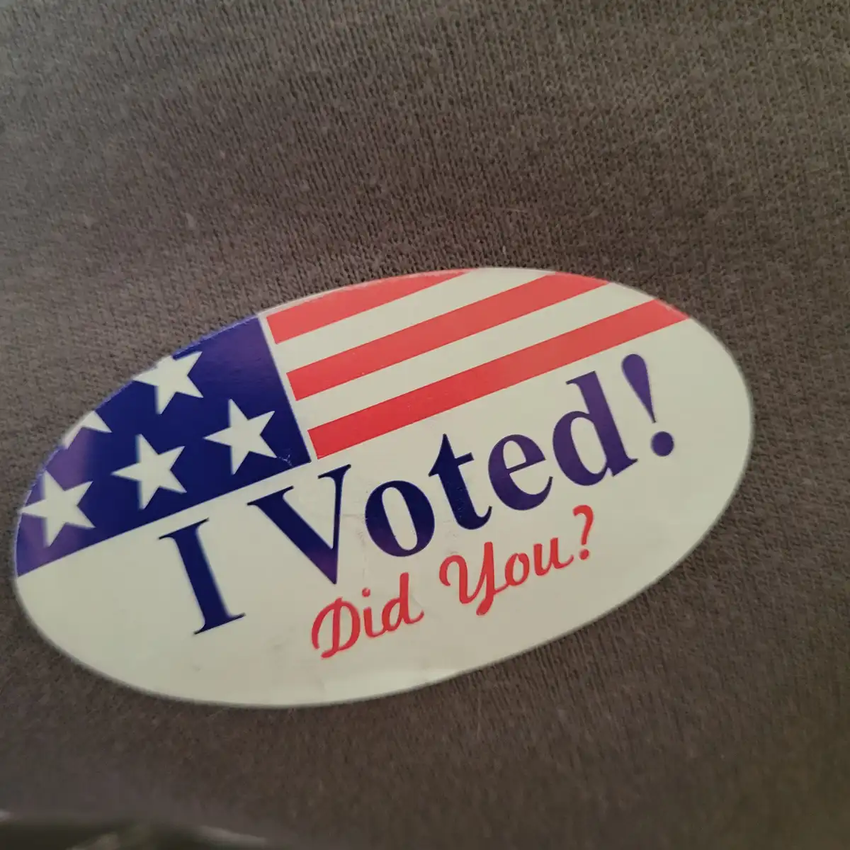 An oval sticker with a U.S. flag design on a gray fabric surface. The sticker features the text 'I Voted!' in large purple…