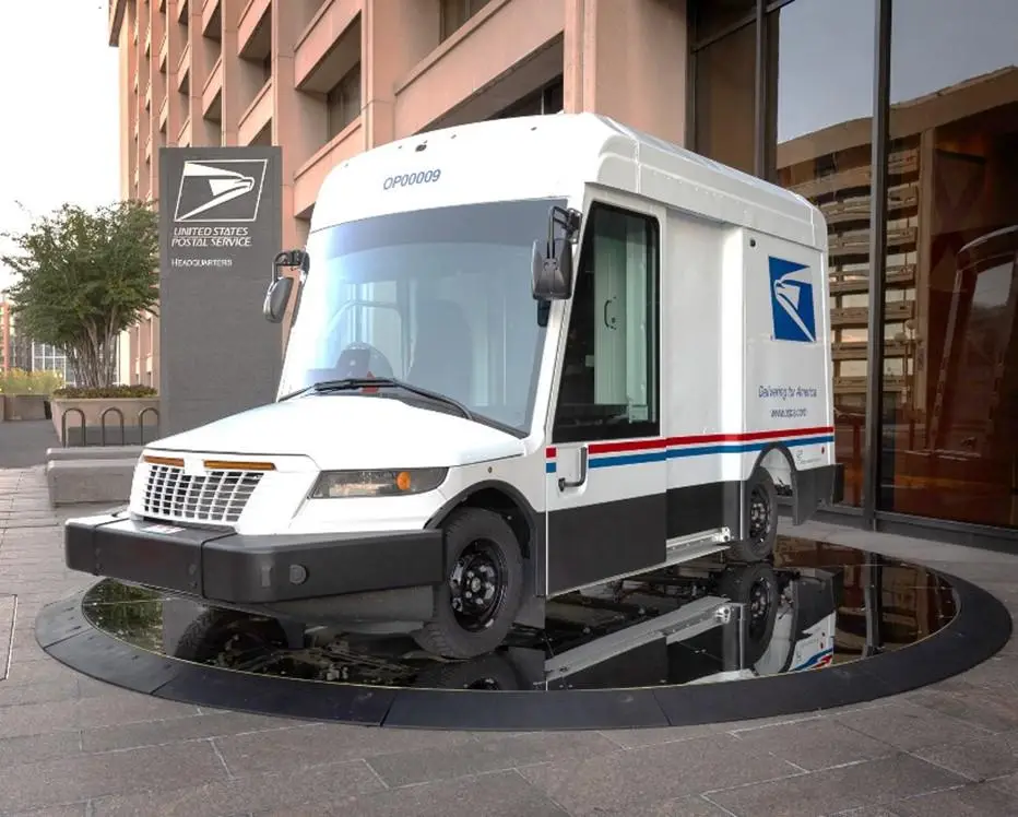 A white United States Postal Service delivery truck is parked on a circular, highly reflective black surface that appears to…