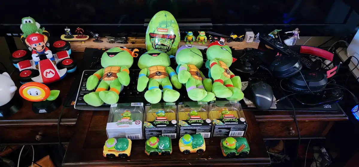 A cluttered desk displays a large collection of Teenage Mutant Ninja Turtles merchandise, including four large plush turtles…