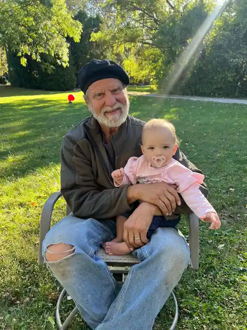 An older man with a white beard and black beret sits outdoors on a lawn chair holding a baby girl in a pink shirt with a…