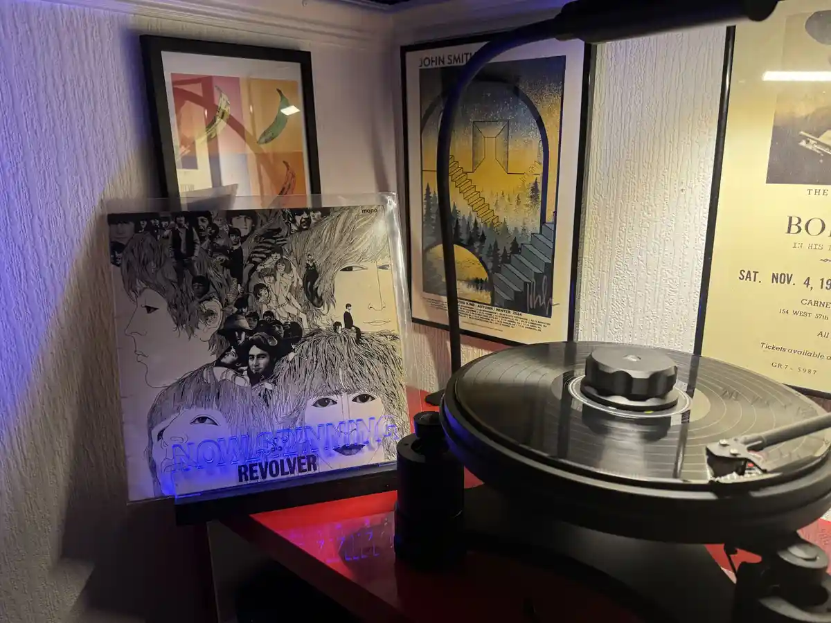 A vinyl record player sits on a red shelf next to a framed Beatles 'Revolver' album cover, which has glowing blue text…