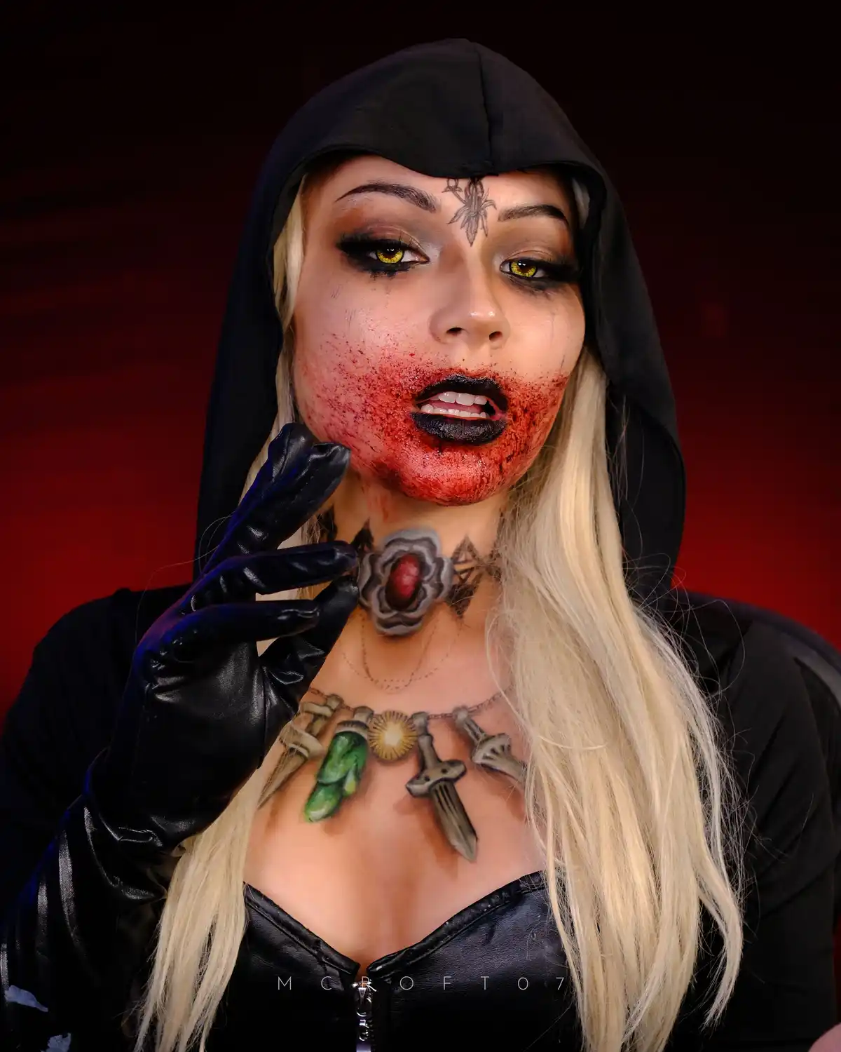 A person with blonde hair and a black hooded outfit poses with dramatic makeup, including yellow eyes, black lipstick, and…