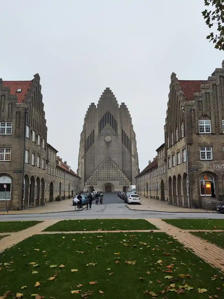 A wide-angle photo shows a large, imposing, modernist church with a stepped, geometric facade, flanked by traditional brick…