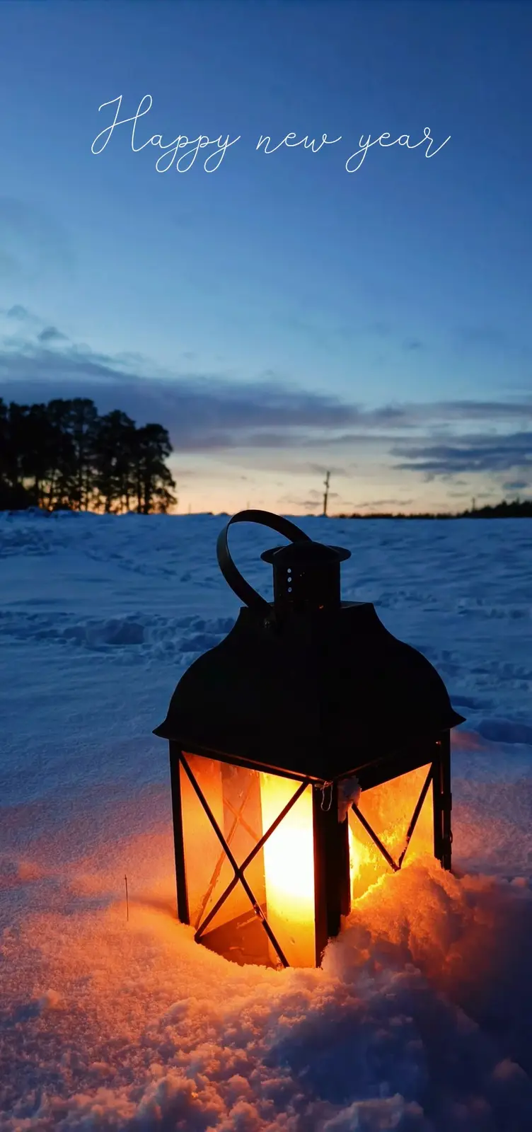 A warmly lit black lantern sits partially buried in snow under a twilight sky, with trees visible in the distance. The…