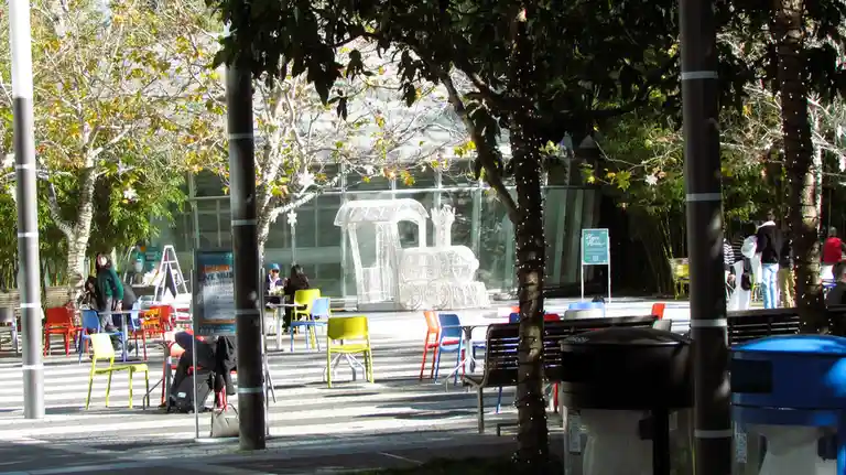 An outdoor plaza with colorful chairs and tables, trees with string lights, and a large white wireframe train sculpture in…