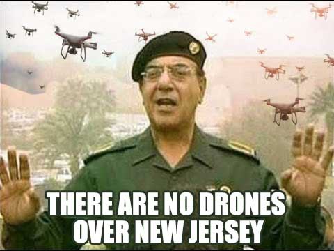 A man in a military uniform with glasses and a beret stands with both hands raised, palms outward, as numerous drones fly…