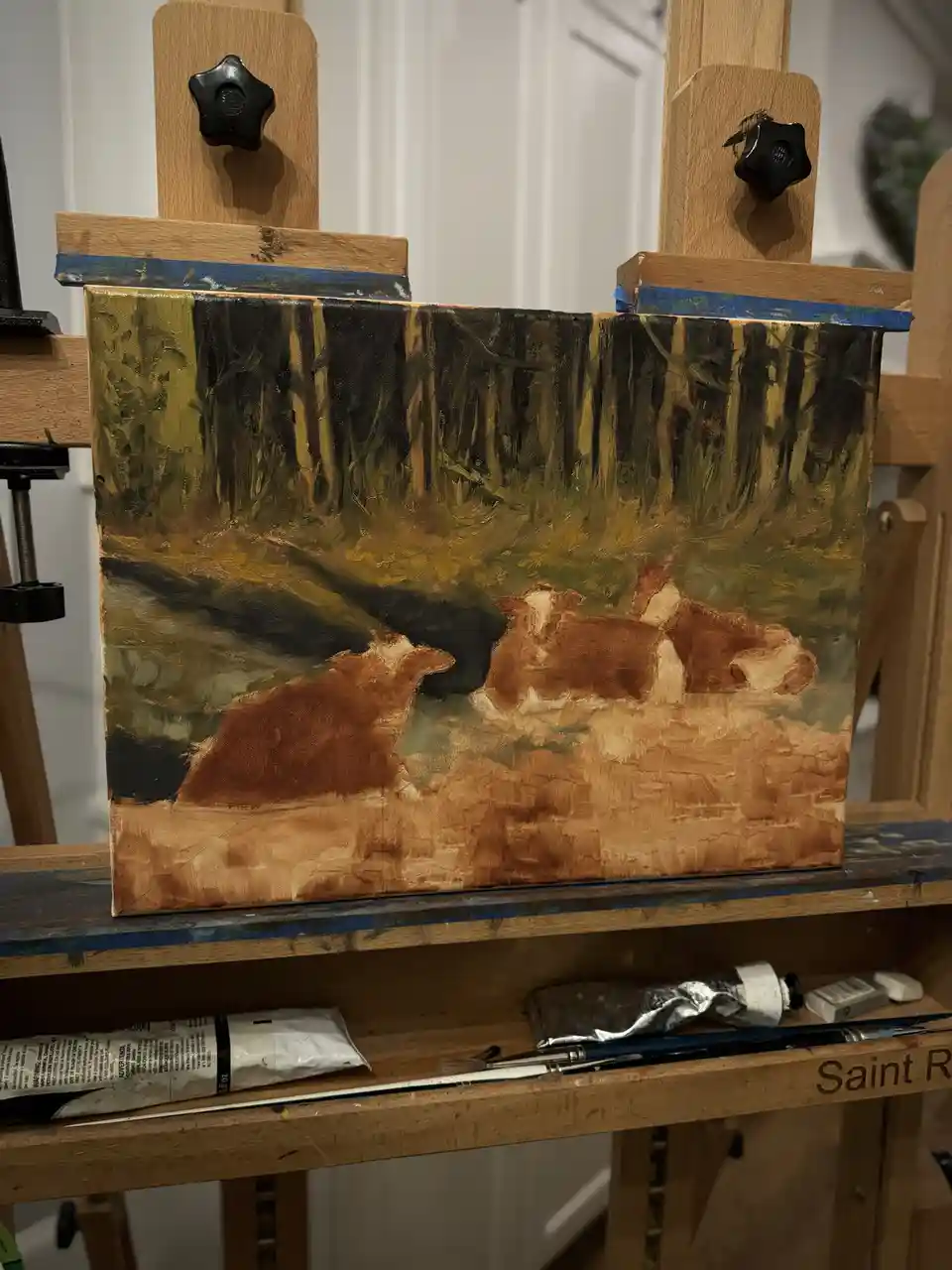 A painting of three brown and white cows resting in a forest clearing is displayed on a wooden easel. The artwork, rendered…