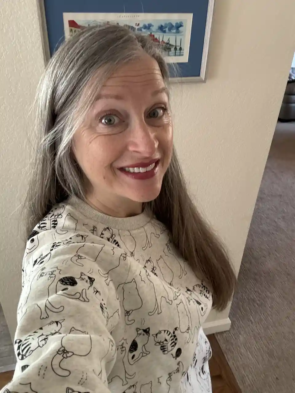 A woman with long gray hair and a wide, slightly startled smile looks at the camera while wearing a light-colored sweatshirt…