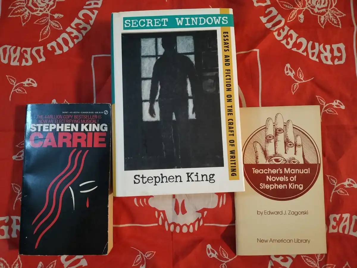 Three Stephen King books are arranged on a red fabric with white skull and rose patterns. The left book is 'Carrie' with a…