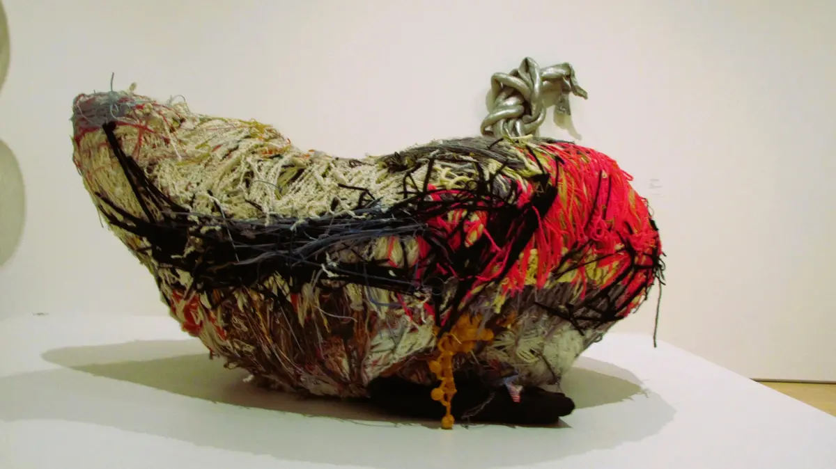 A large, abstract sculpture resembling a distorted, elongated heart or boot made from tangled yarns, threads, and fabric…