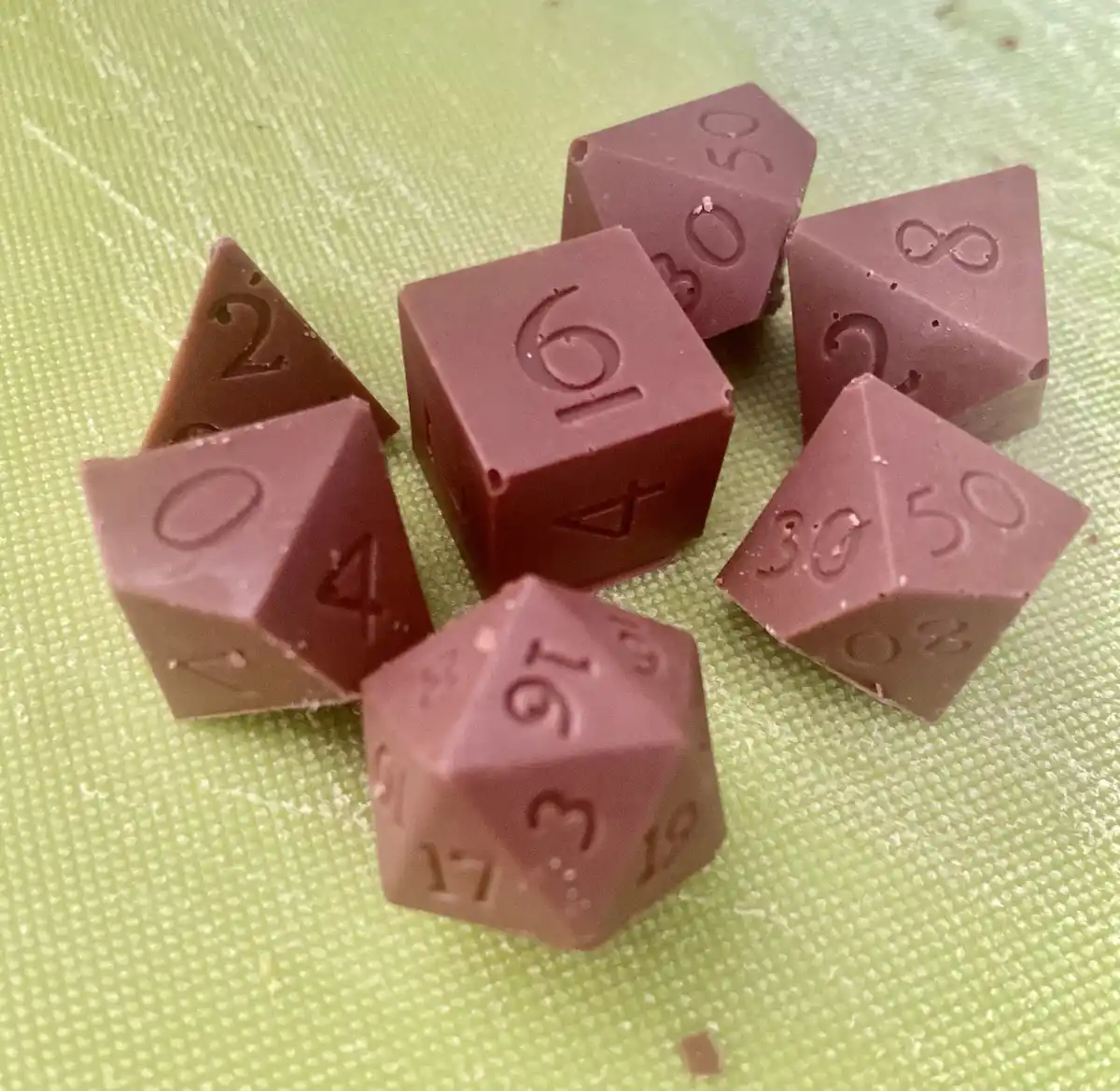 Several brown, chocolate-like dice shaped like polyhedral dice used in Dungeons & Dragons are scattered on a light green…