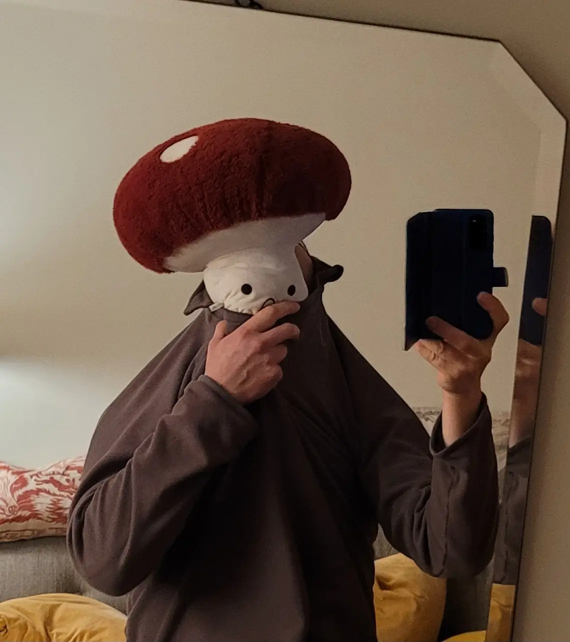 A person wearing a large, plush red-and-white mushroom hat takes a mirror selfie while holding a phone in one hand and…