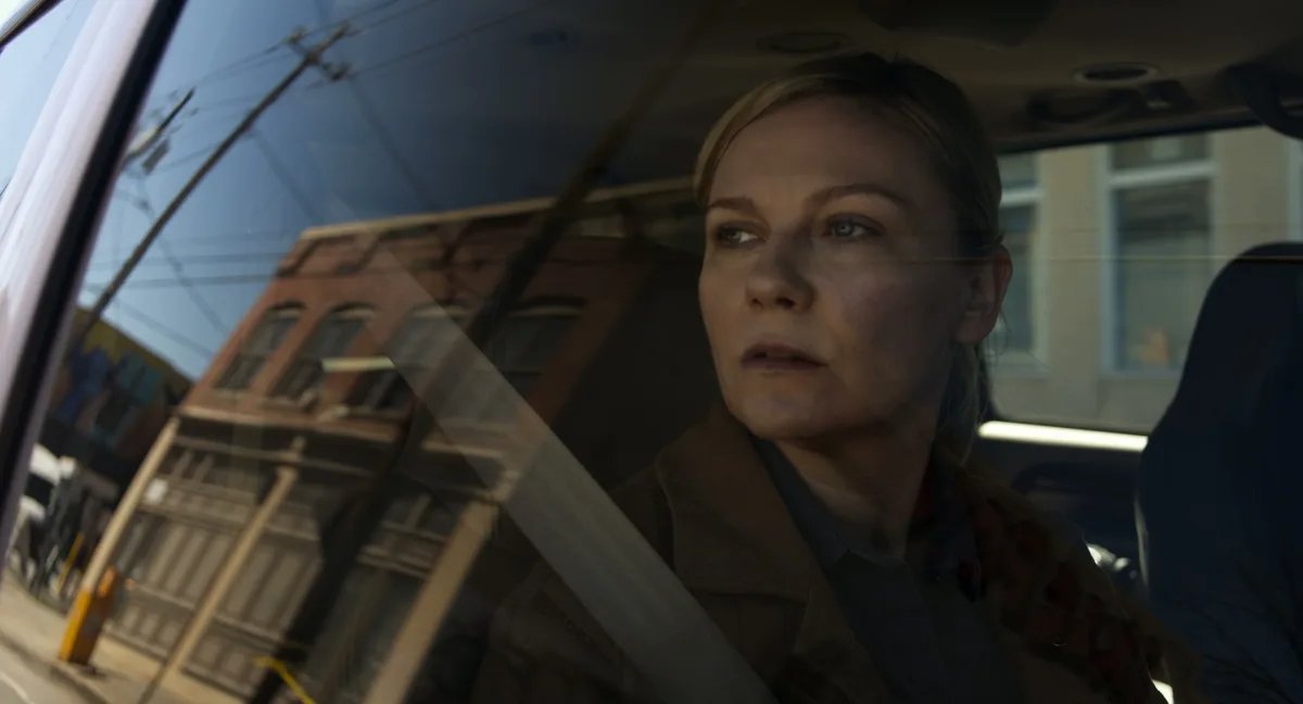 A woman with blonde hair sits in a car, looking out the window with a somber expression. The car's interior and a blurred…