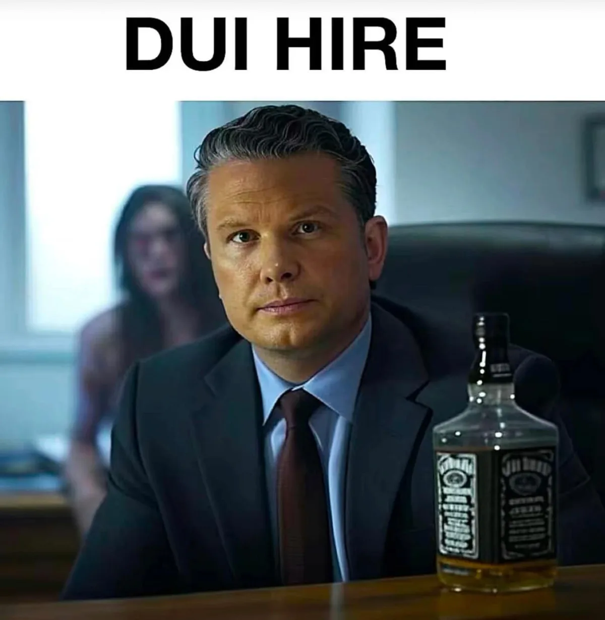 A man in a suit sits at a desk with a bottle of Jack Daniels whiskey in front of him, looking serious. Behind him, out of…