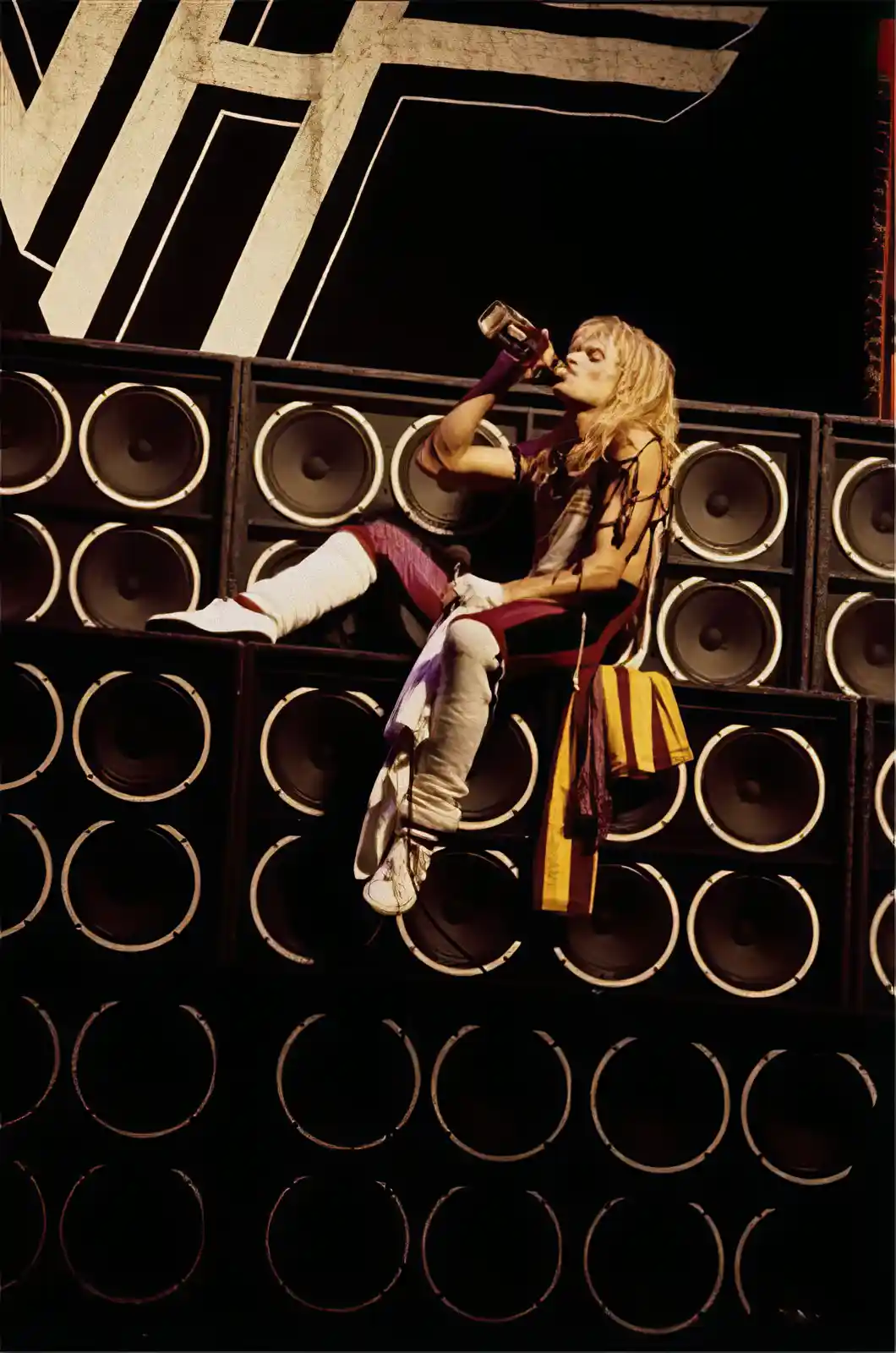 A person with long blonde hair, wearing a strappy top and white boots, is sitting atop a large wall of vintage speakers…