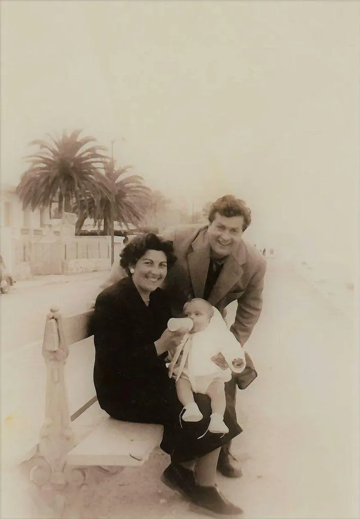 A sepia-toned vintage photo shows a smiling couple posing on a bench with a baby. The woman sits holding the baby, who is…