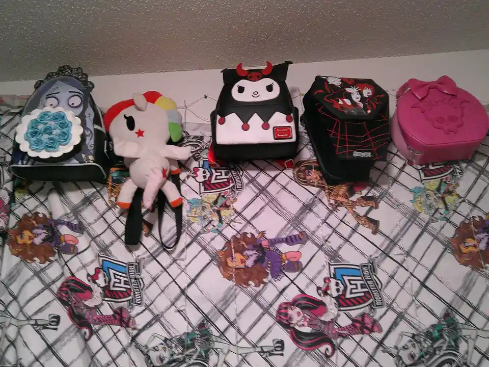 Five themed bags and a plush toy are arranged on a Monster High-patterned surface. From left to right: a blue skull-faced…