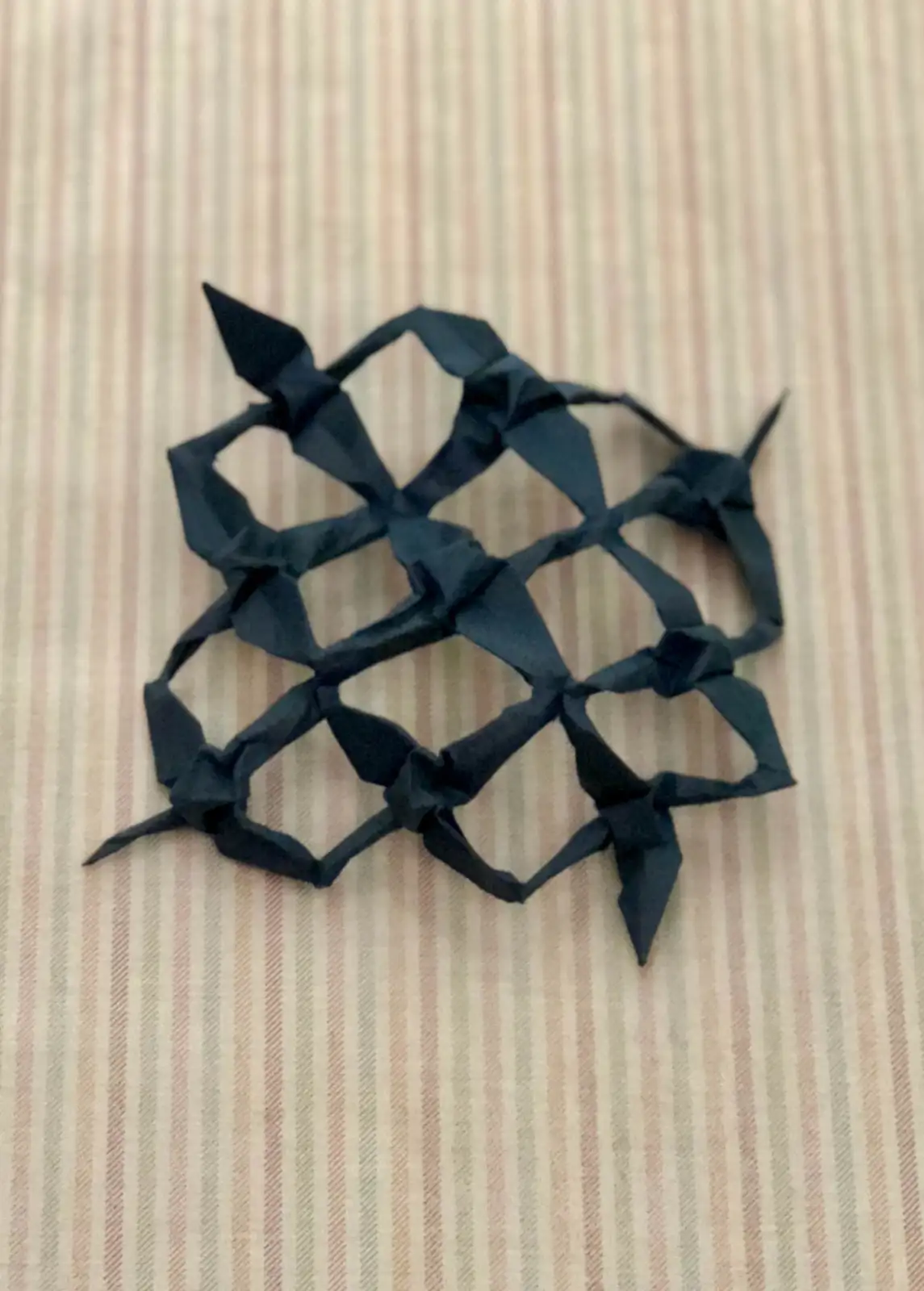 A black origami creation with multiple pointed legs and a web-like structure rests on a striped fabric background. The…
